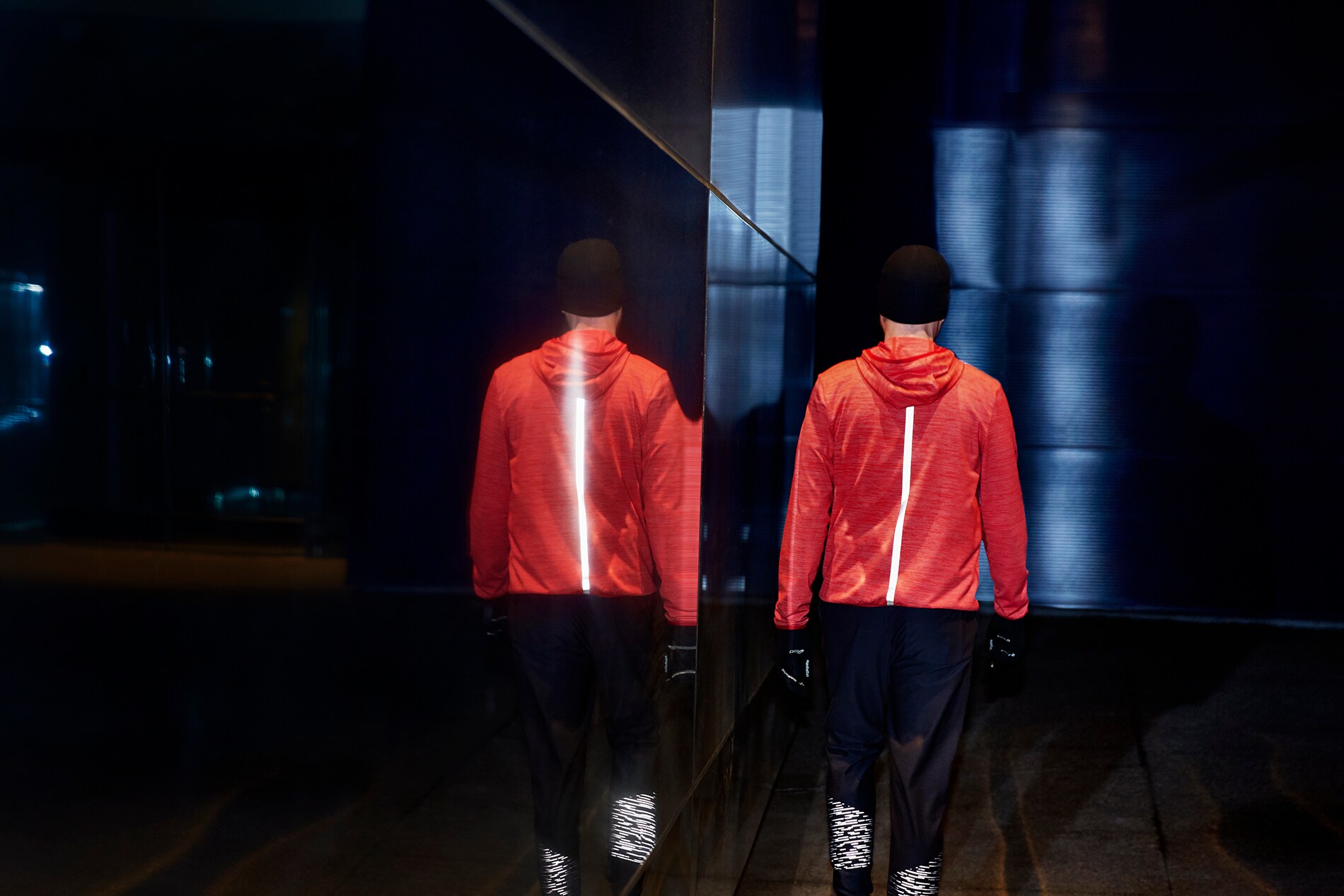 Strisce di materiale riflettente elastico nero illuminato sul retro di una giacca da corsa di un uomo che cammina accanto a una parete, di specchi di notte
