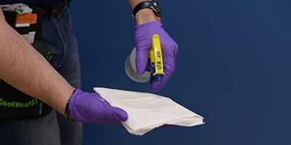 Persona con guanti chirurgici blu che spruzza su un foglio di carta assorbente lo spray consigliato da 3M per aumentare l'adesione delle pellicole
