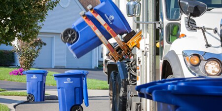 Un bidone blu per il riciclo viene sollevato su un camion per la raccolta differenziata.
