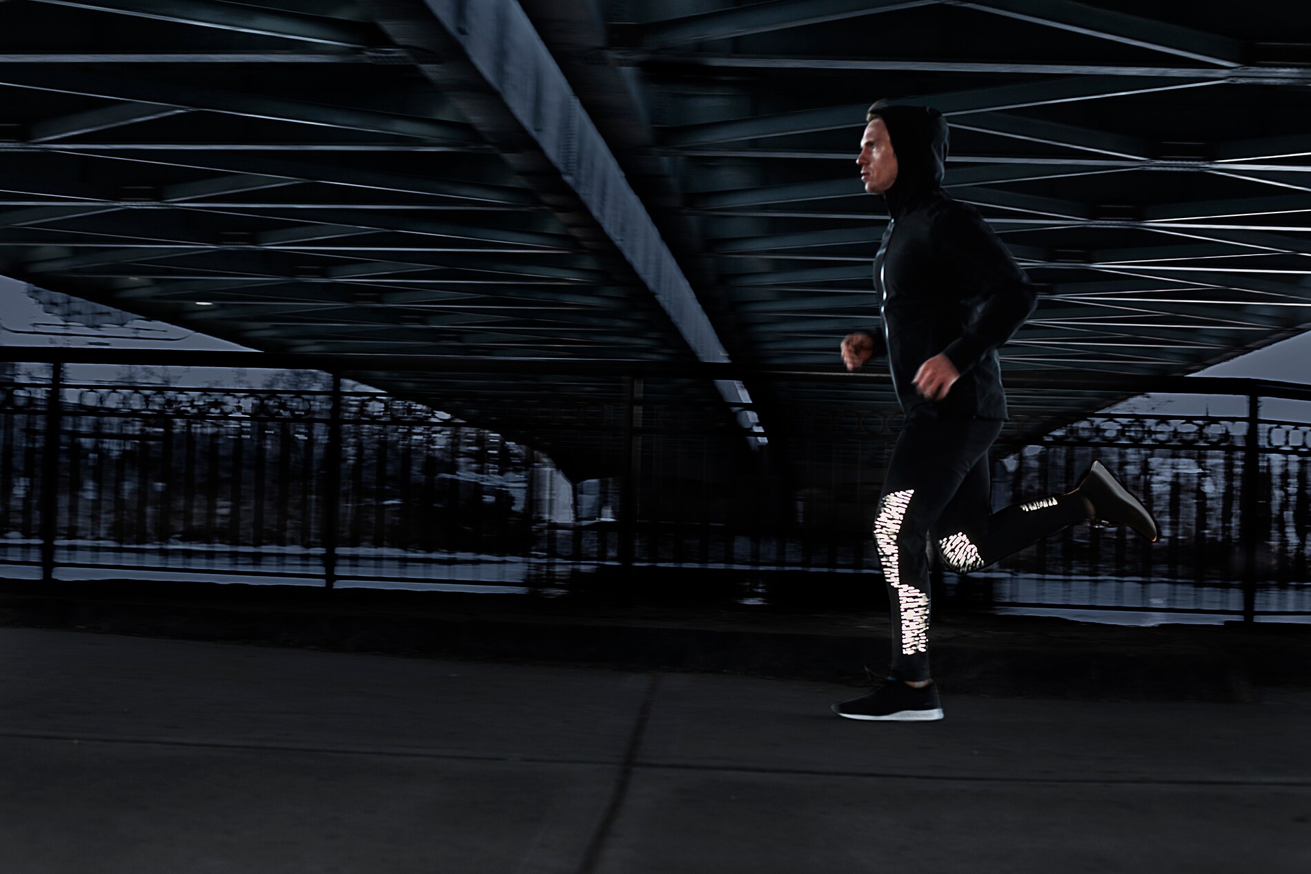 Un uomo e una donna sotto un ponte cittadino al crepuscolo che indossano leggings da corsa illuminati, realizzati con materiale riflettente elastico nero 3M Scotchlite