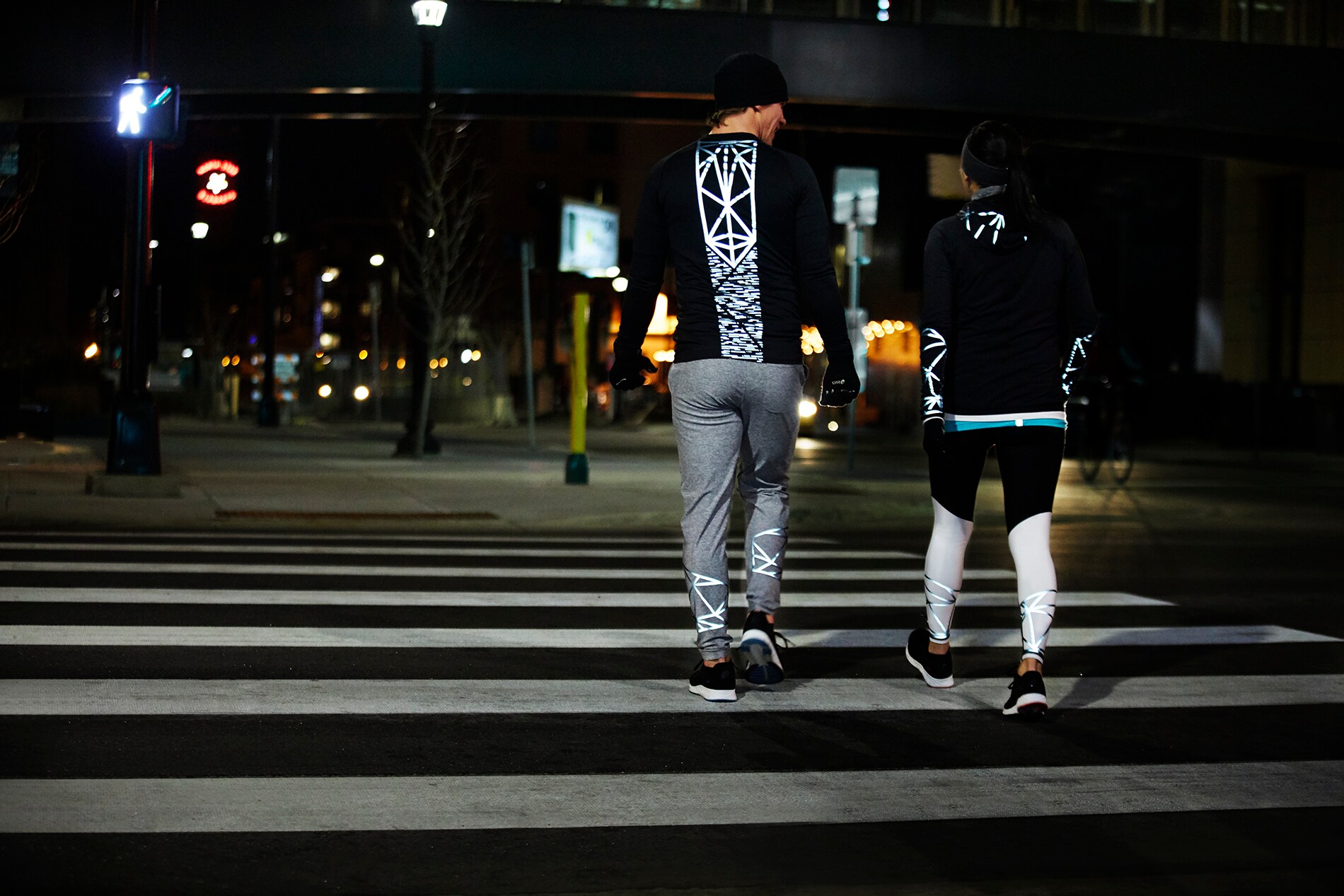 Un uomo e una donna che corrono, durante un tour notturno della città, e indossano abbigliamento tecnico illuminato, realizzato con materiale riflettente elastico nero 3M Scotchlite
