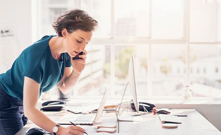 Donna al telefono in piedi sulla scrivania a prendere appunti in un ufficio bianco luminoso, soleggiato e con grandi finestre
