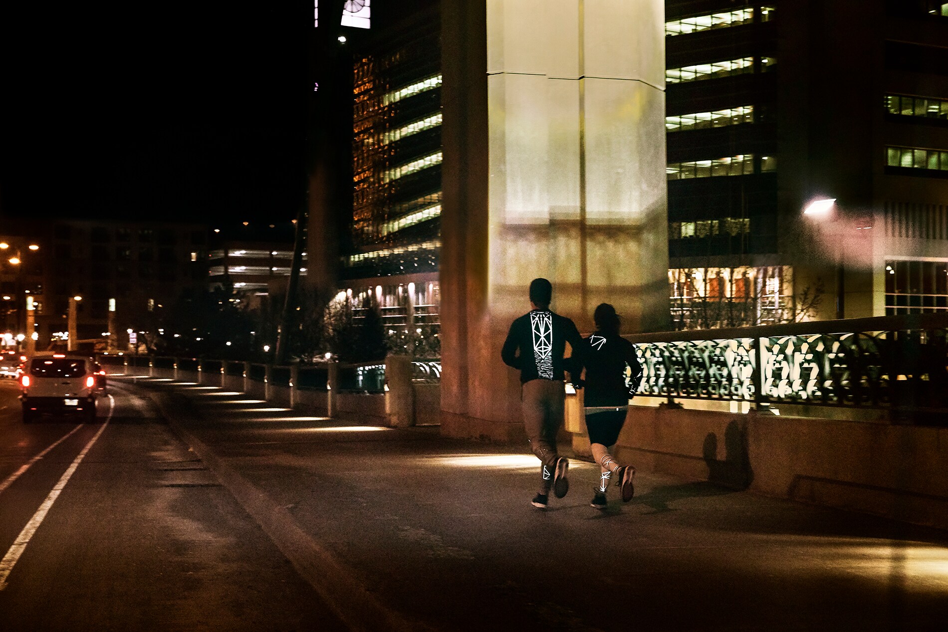 Un uomo e una donna che attraversano di corsa un ponte in città e indossano abbigliamento tecnico per la corsa illuminato, realizzato con materiale riflettente elastico nero 3M Scotchlite