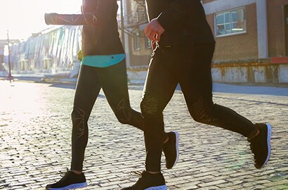 Un uomo e una donna che corrono su una strada di ciottoli al tramonto e indossano leggings con materiale riflettente elastico nero 3M Scotchlite