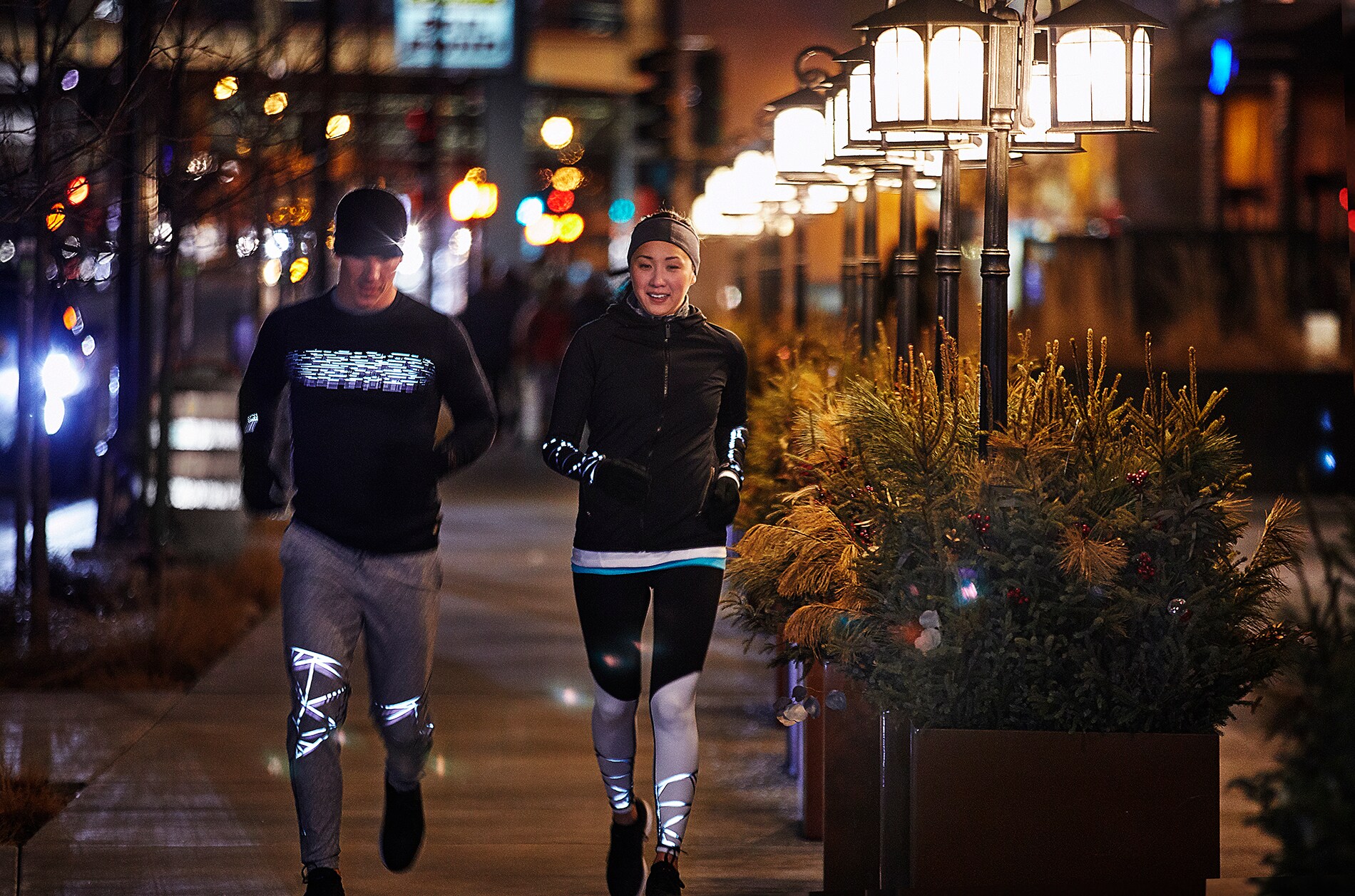 Un uomo e una donna che corrono sotto le luci della città e indossano abbigliamento tecnico per la corsa illuminato, realizzato con materiale riflettente elastico nero 3M Scotchlite